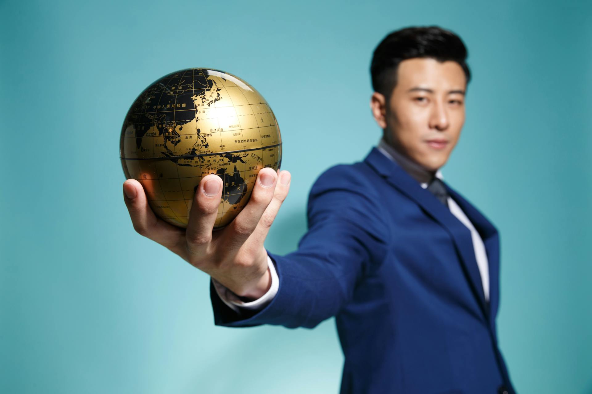 Asian businessman in blue suit confidently holding a golden globe against a blue background.