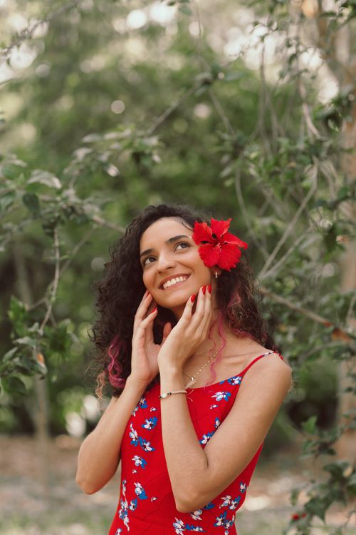 Free Portrait of a smiling woman outdoors with a flower in her hair, wearing a red floral dress. Stock Photo