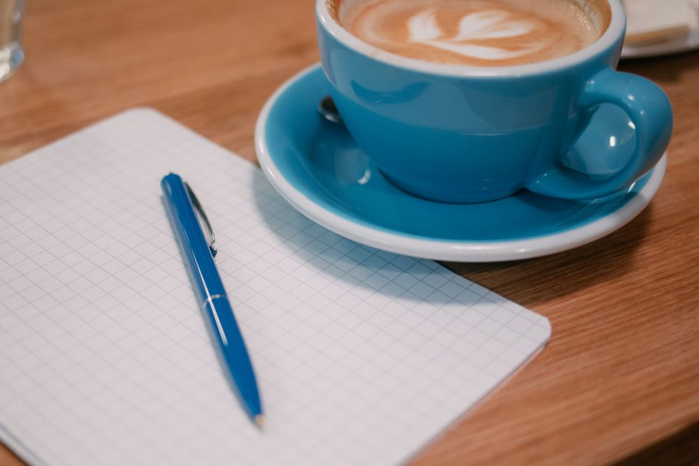 Ceașcă de cappuccino cu desen în spumă, lângă un carnet cu pătrățele și un pix, pe lemn