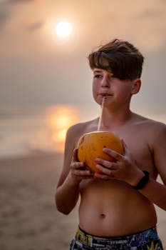 Teenager drinks coconut water at sunset on a sandy beach, enjoying tropical vibes.