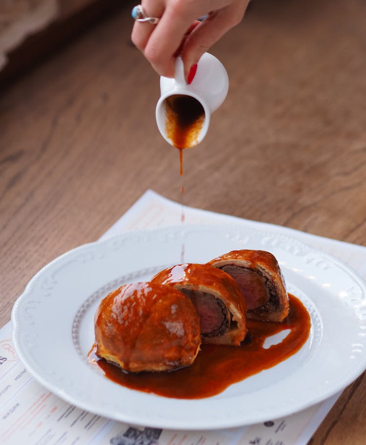 A Hand Pouring Brown Sauce On White Ceramic Plate