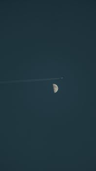 A serene night sky with an airplane silhouetted against a half moon, captured in a vertical shot.