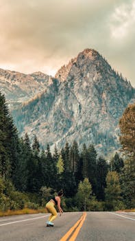 A woman skateboards down a highway surrounded by forest and mountains under a cloudy sky.