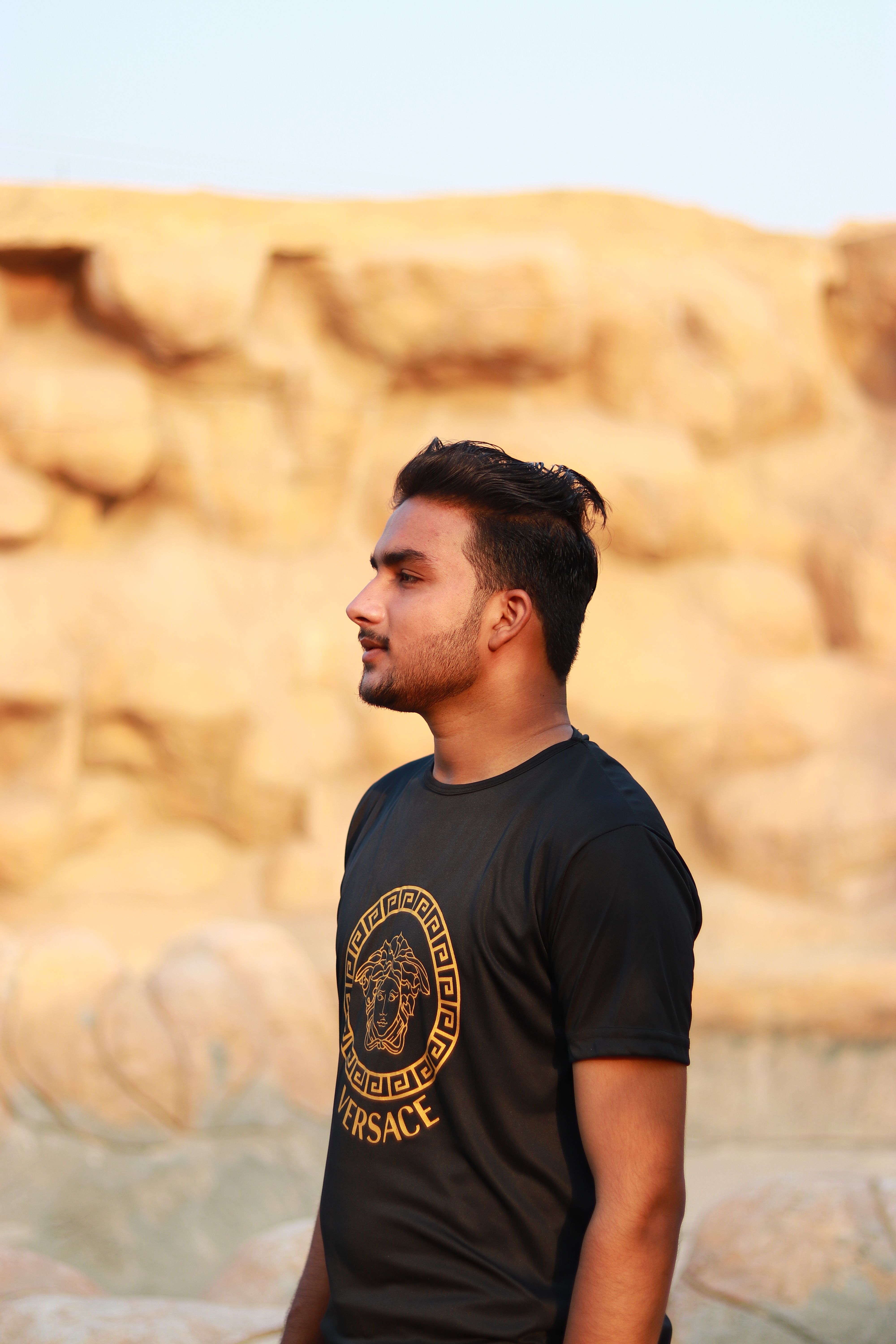 Man standing outdoors against a nature backdrop, wearing a black T-shirt with gold design.
