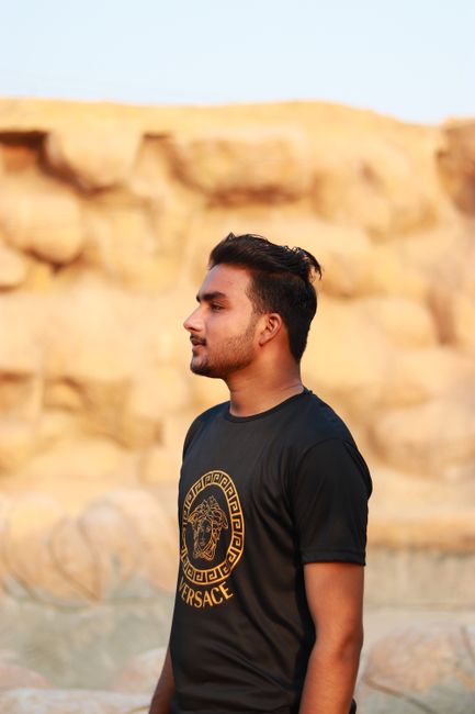 Man standing outdoors against a nature backdrop, wearing a black T-shirt with gold design.