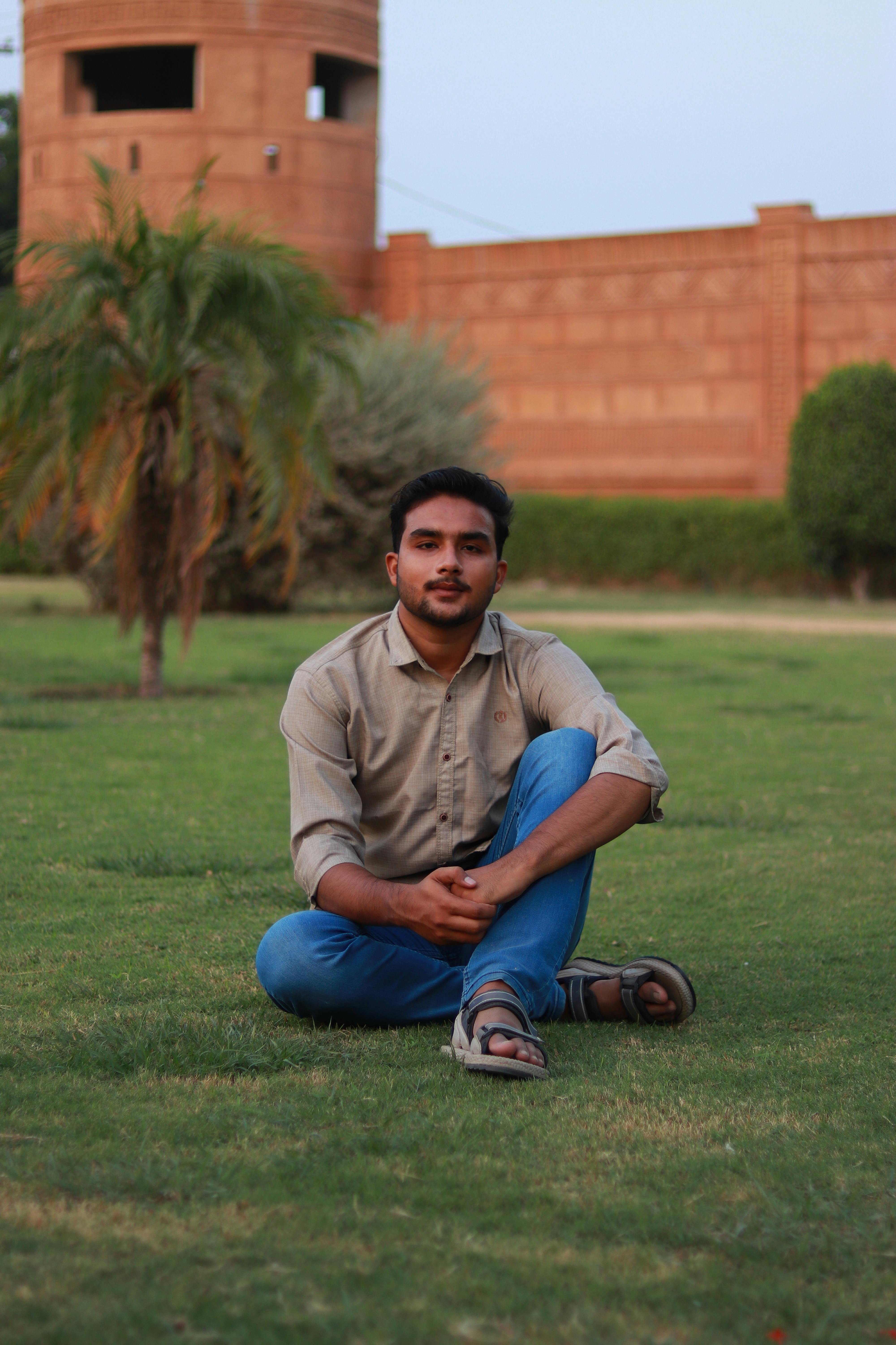 A young man sits cross-legged on a sunny green lawn with a brick structure in the background.