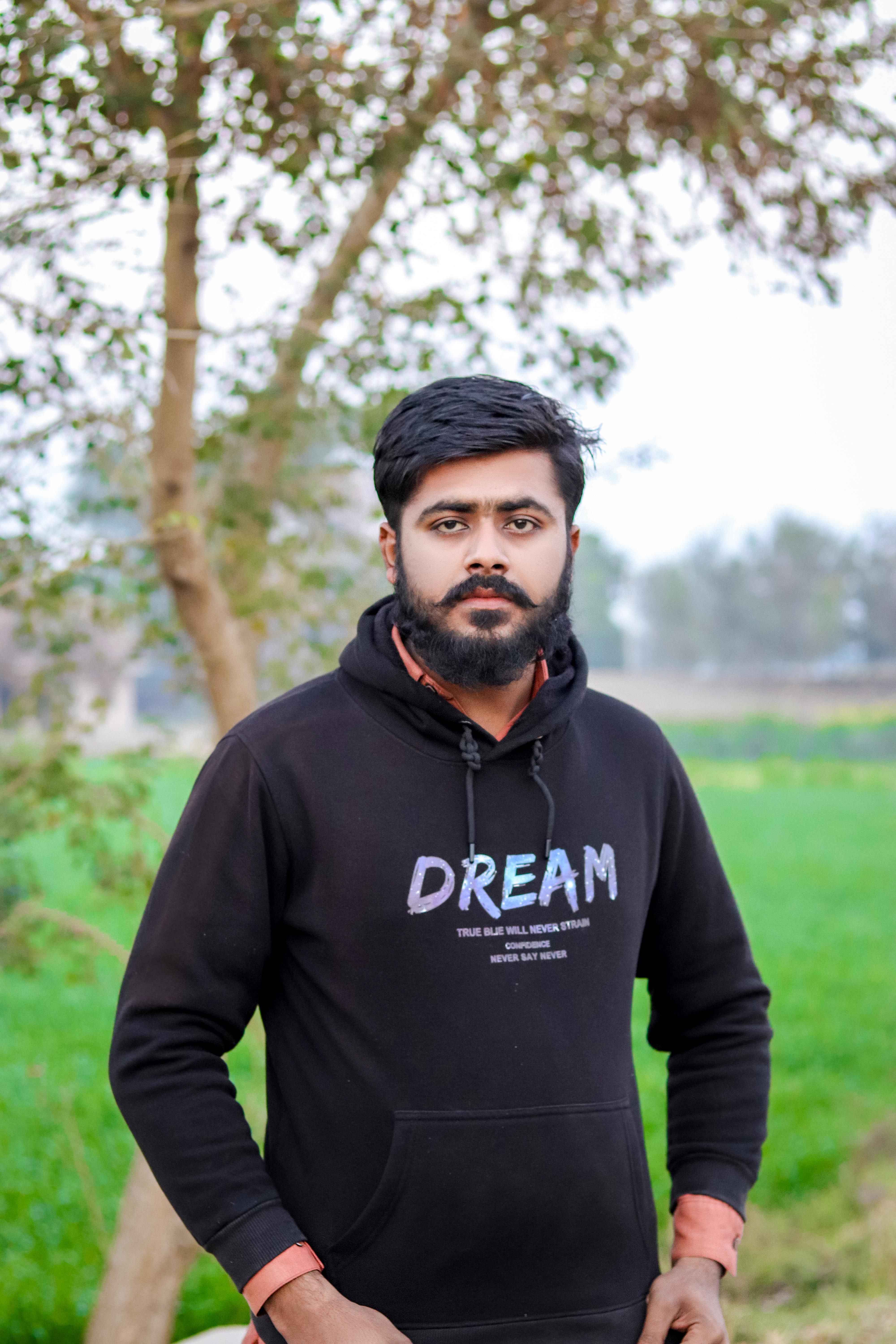 Portrait of a young man with a beard wearing a hoodie outdoors, standing by trees.