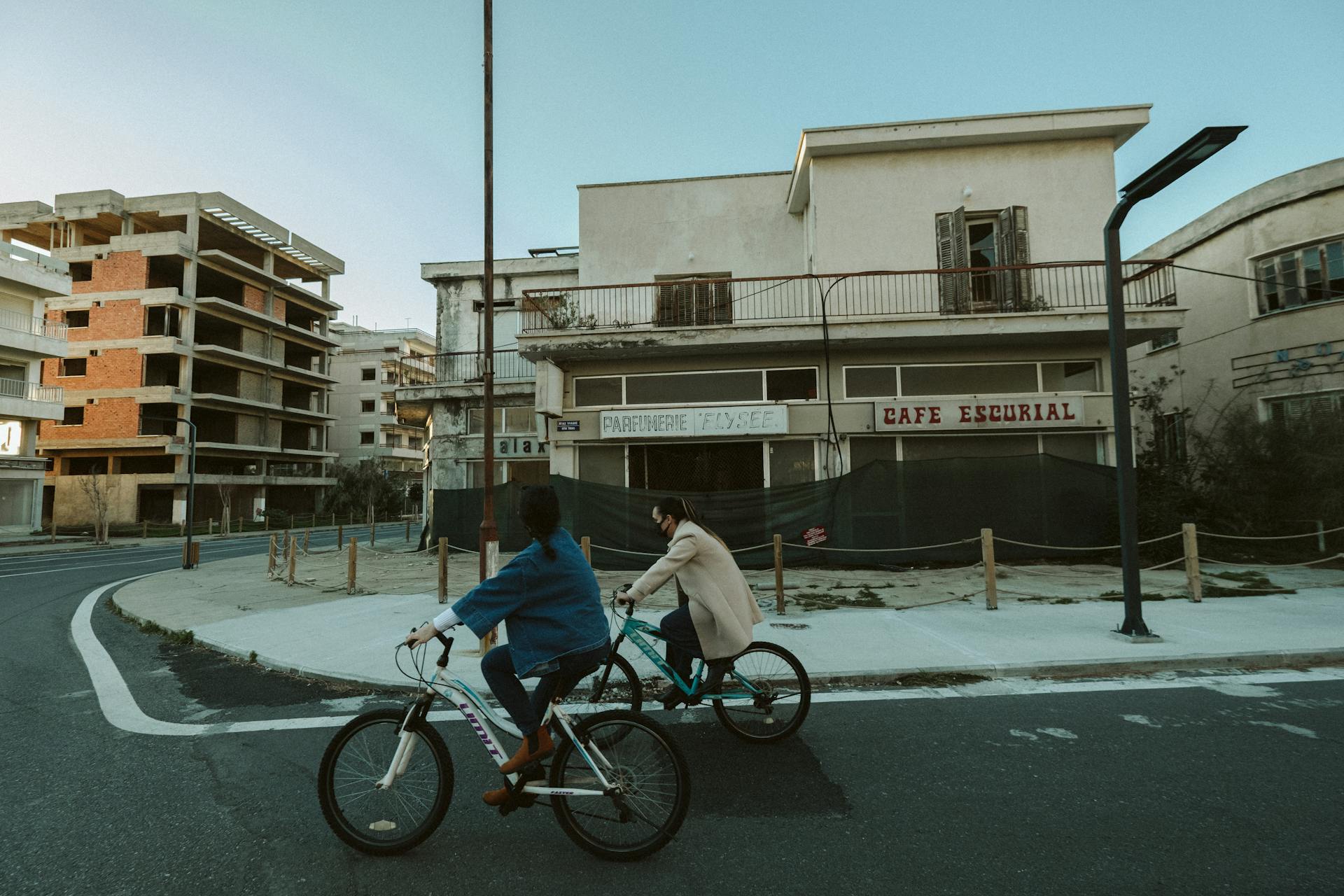 Two cyclists riding past weathered, abandoned buildings in a quiet, urban street setting.