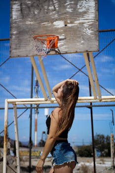 Woman in casual outfit posing outdoors by an old basketball hoop on a sunny day.