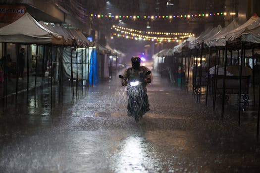 Motorcycle rider braves heavy rain in Bangkok street lined with tents and string lights at night.