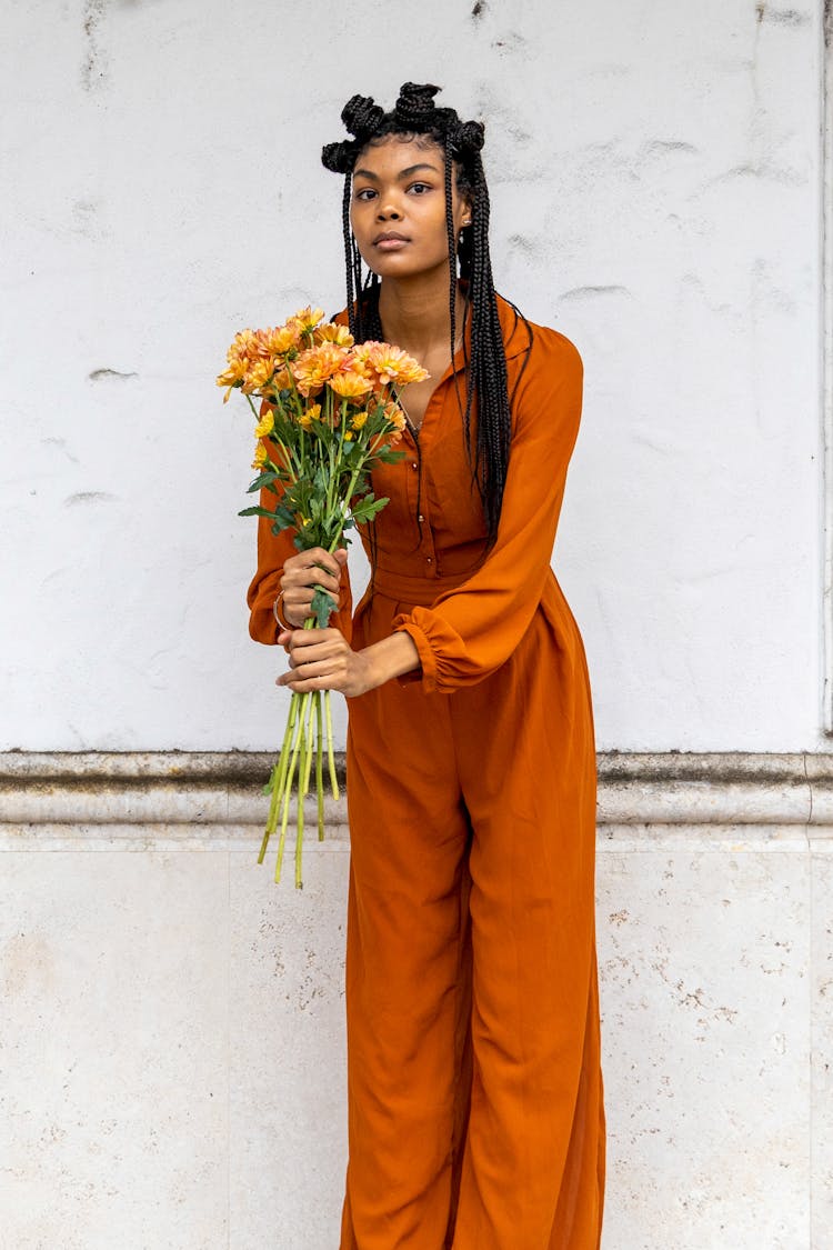 Woman In Orange Jumpsuit Holding Stems Of Flower While Looking At The Camera