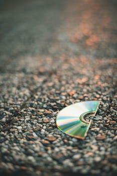 A broken CD lies on gravel reflecting sunlight, conveying obsolescence.