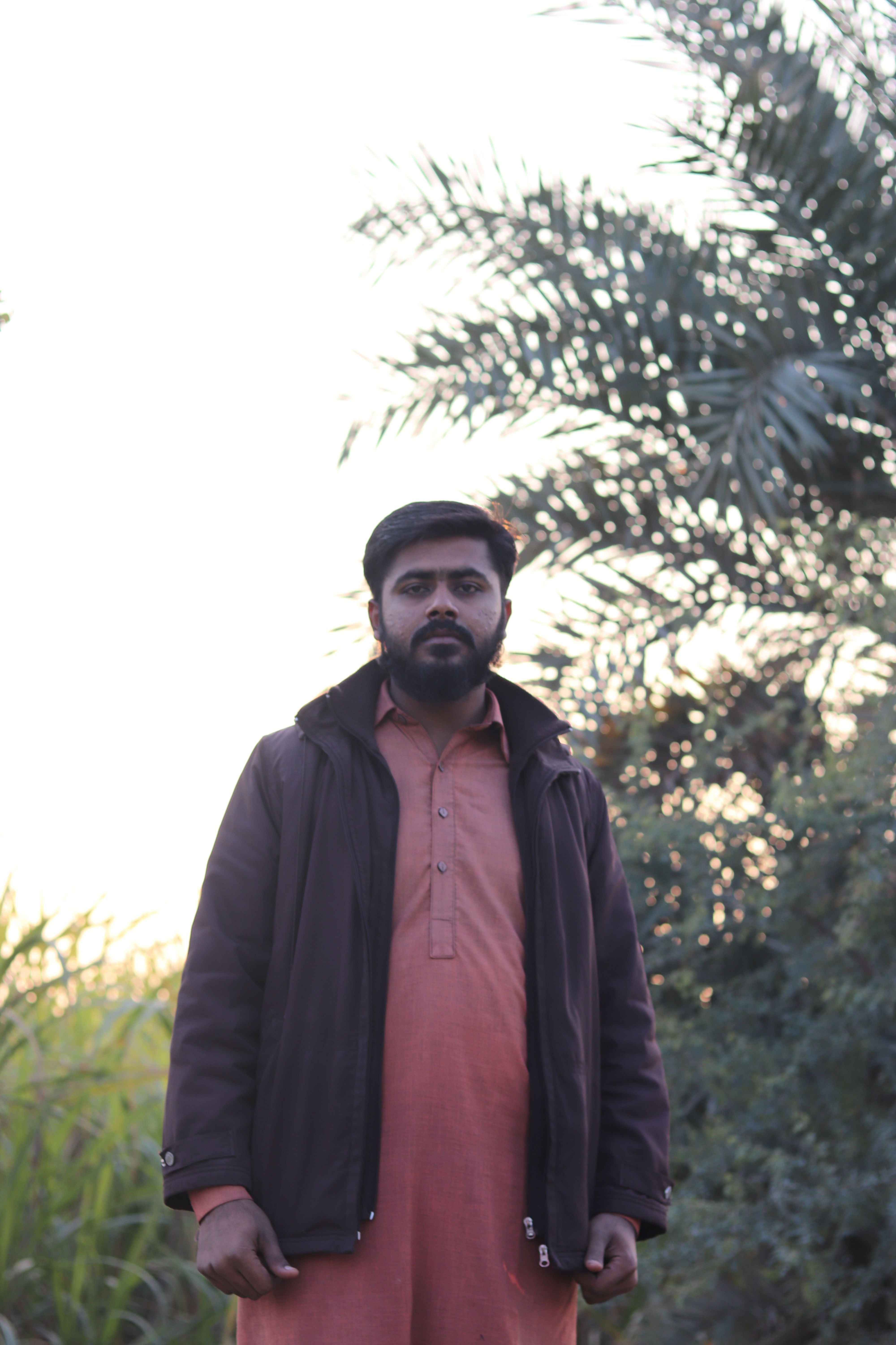 A man in traditional attire standing outdoors by greenery, captured at sunset.