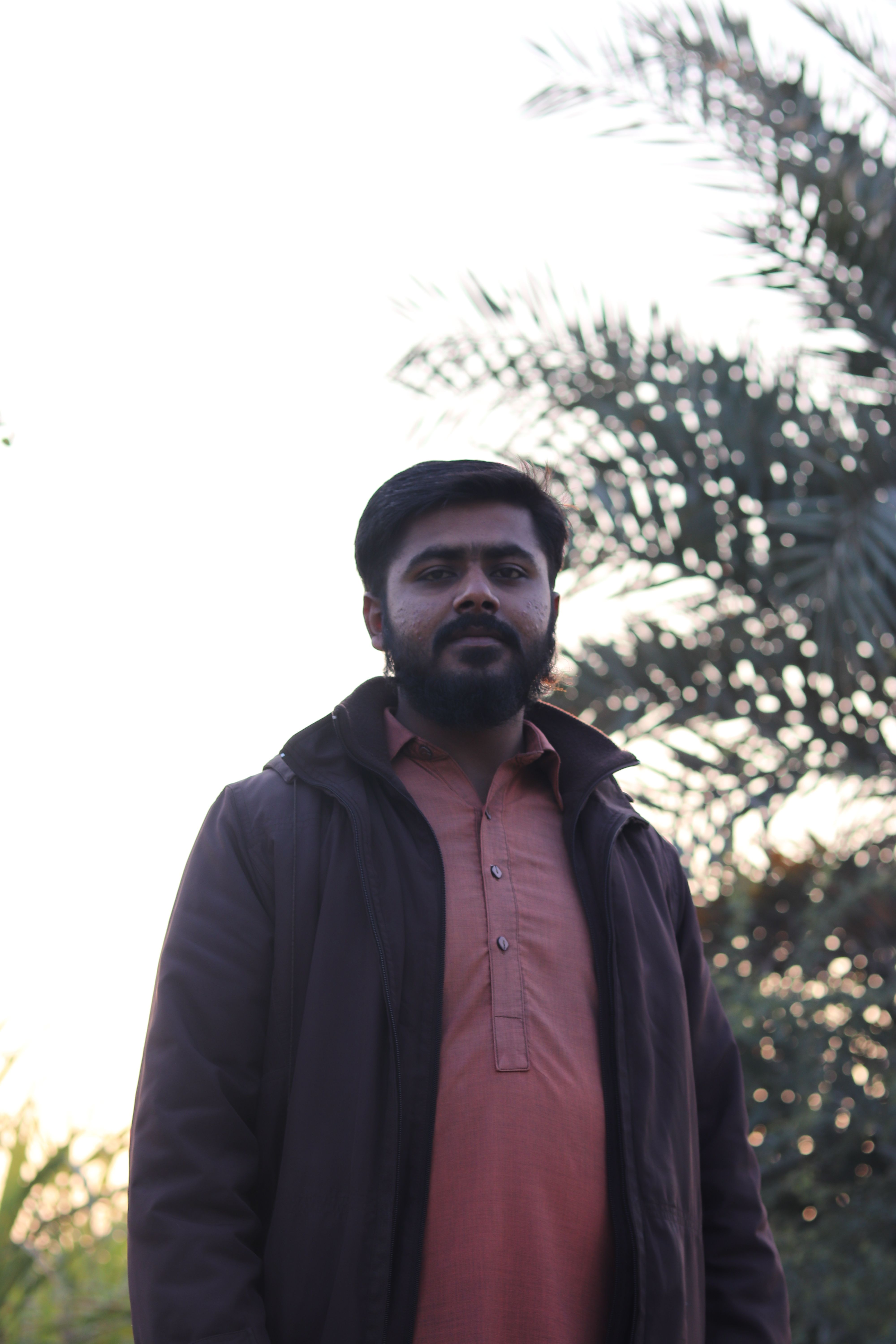 A man with a beard standing outdoors with natural light behind him, wearing a jacket.
