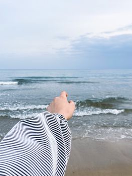 A hand in a striped sleeve extends towards a calm sea, under a cloudy sky.