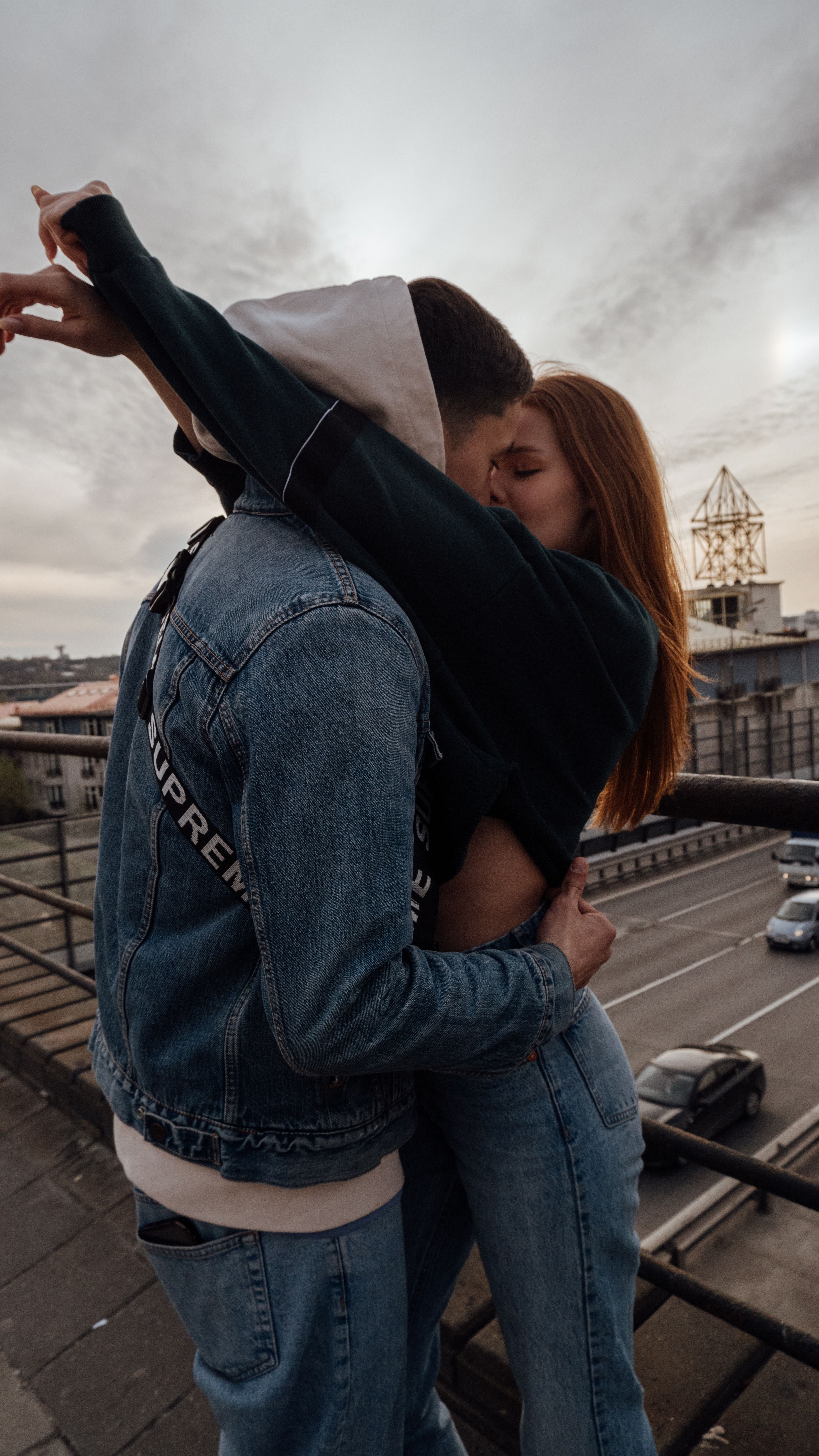 A young couple shares a romantic embrace and kiss on a city rooftop at dusk.