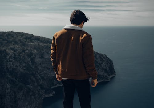 Back View of a Man Wearing Brown Jacket at the Edge of a Cliff