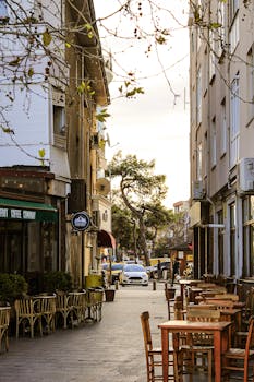 A cozy street in Çanakkale featuring outdoor cafes, tables, and buildings bathed in warm light.