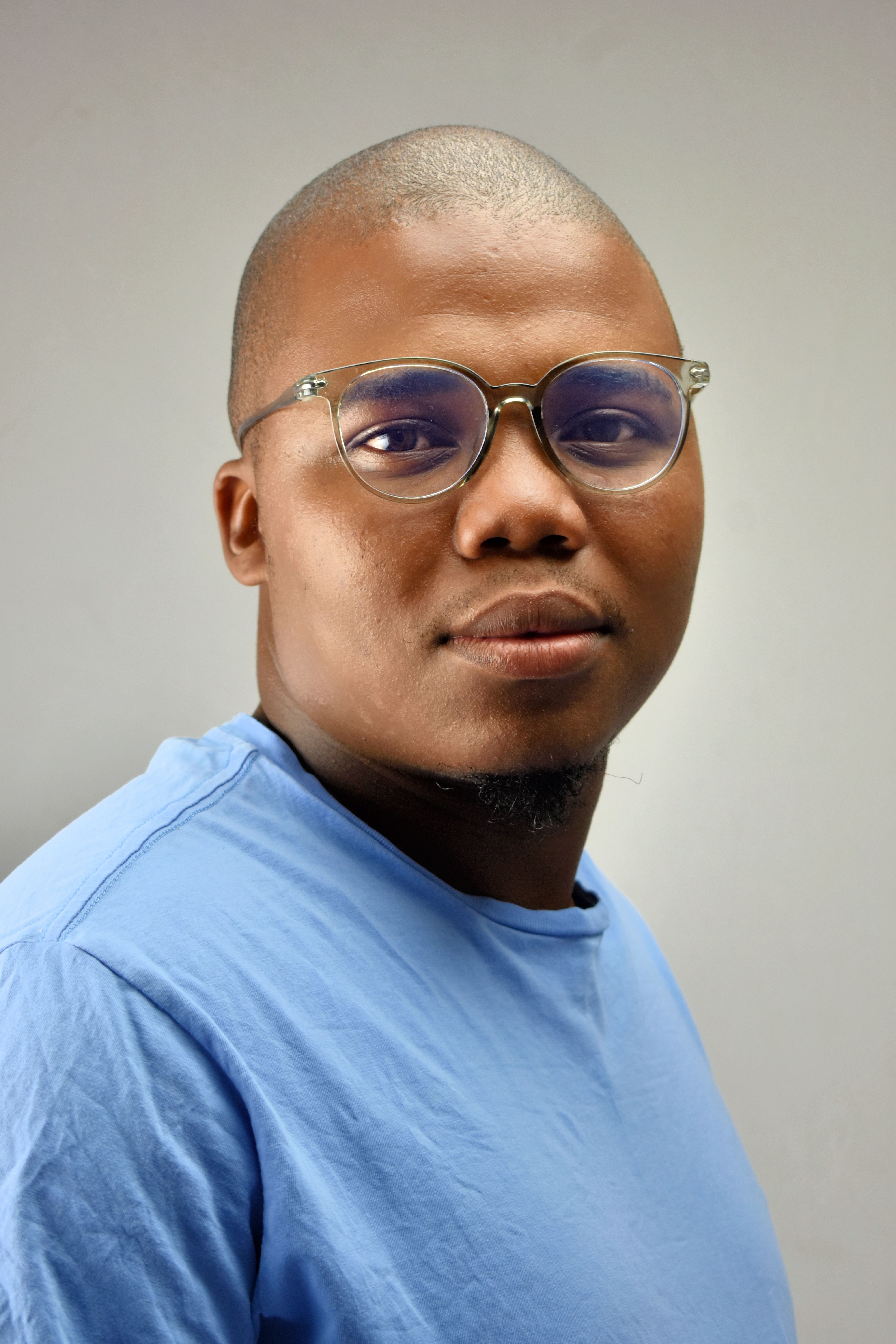 A confident young man wearing glasses and a blue shirt against a neutral background.
