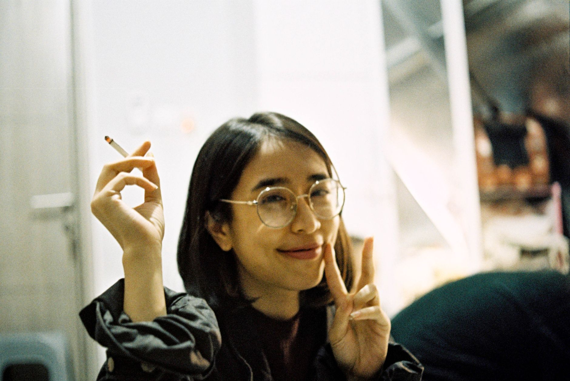 Cheerful young woman indoors with glasses, holding a cigarette and making a peace sign.