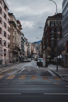 City street with parked cars and buildings under a moody, cloudy sky.