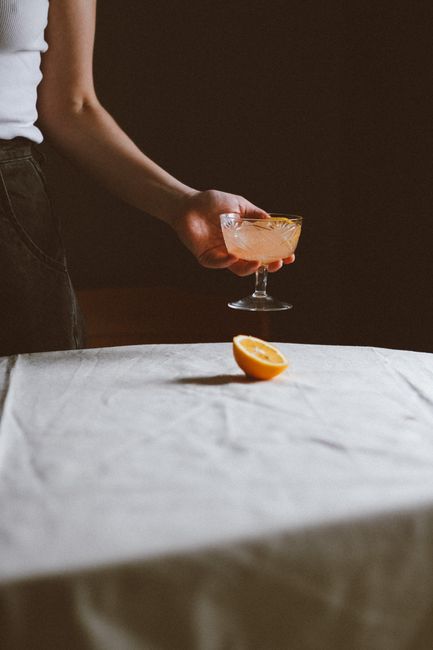 A woman holds a citrus cocktail in a vintage glass on a draped table with orange half.