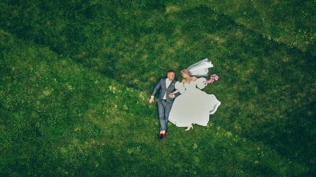 Beautiful aerial view of a newlywed couple lying on green grass, bride in wedding dress, groom in suit, sharing a moment of love.