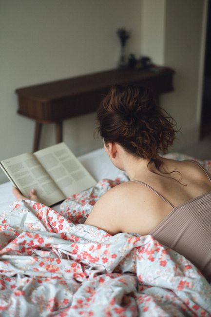 A woman is comfortably reading in bed with floral sheets, creating a warm and serene atmosphere.