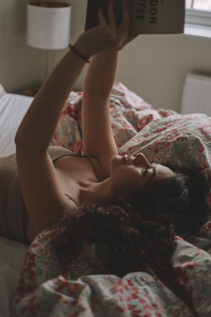 A young woman lies in bed, reading a book in a cozy, softly lit bedroom.