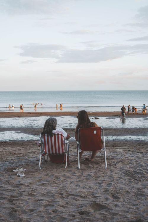 Free Two people enjoy a peaceful day at the beach, sitting on chairs by the sea with a view of the shoreline. Stock Photo