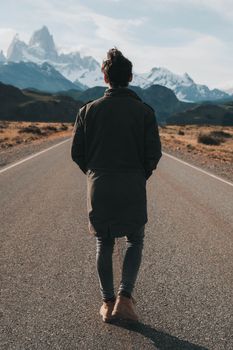 Back view of a man walking on a deserted road towards snowy mountains under a clear sky.