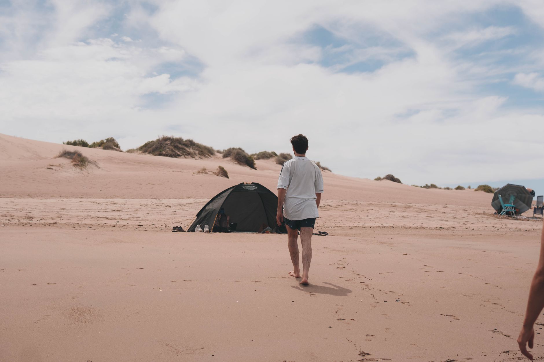 A man walks on a sandy beach towards a tent under a cloudy sky, evoking a sense of adventure.