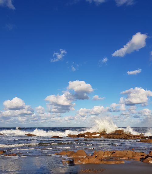 Free Waves crash against rocky shore under a vibrant blue sky, capturing the essence of a summer day. Stock Photo