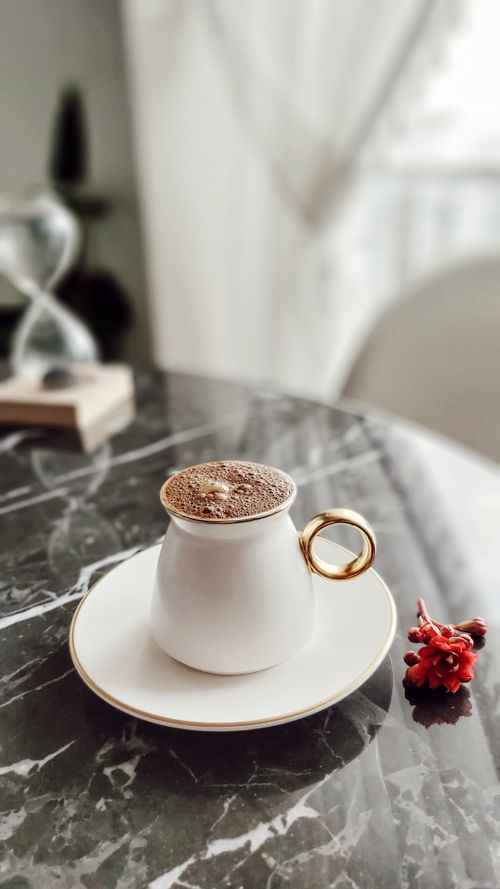 Free Close-up of a white coffee cup with gold handle on a marble table with a red flower accent. Stock Photo