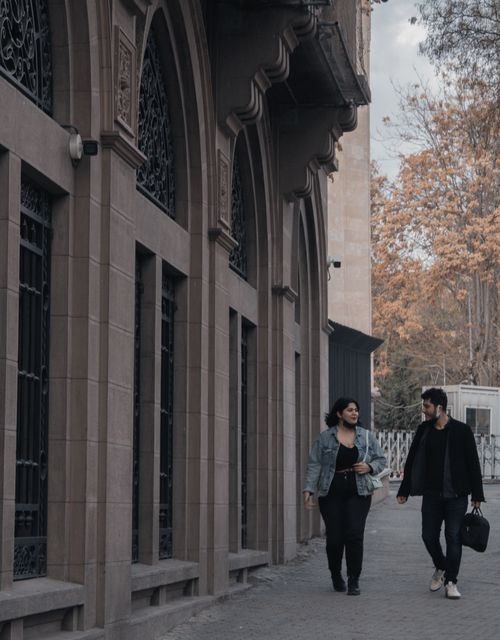 Free Two adults walking and talking on a sidewalk by an arched stone building during autumn. Stock Photo