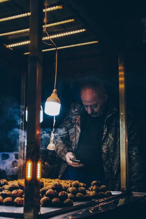 Man in Black Winter Jacket Busy Using His Phone while Standing in Front of a Food Stall