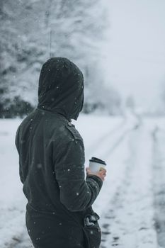 A person in a hoodie holds a coffee cup while standing on a snow-covered road during winter snowing.