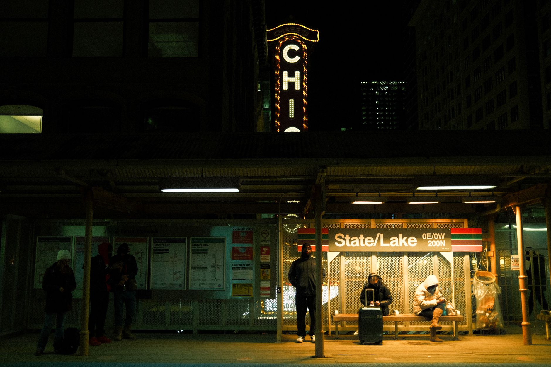 Transit passengers waiting at Chicago's State/Lake station platform at night, under neon lights.