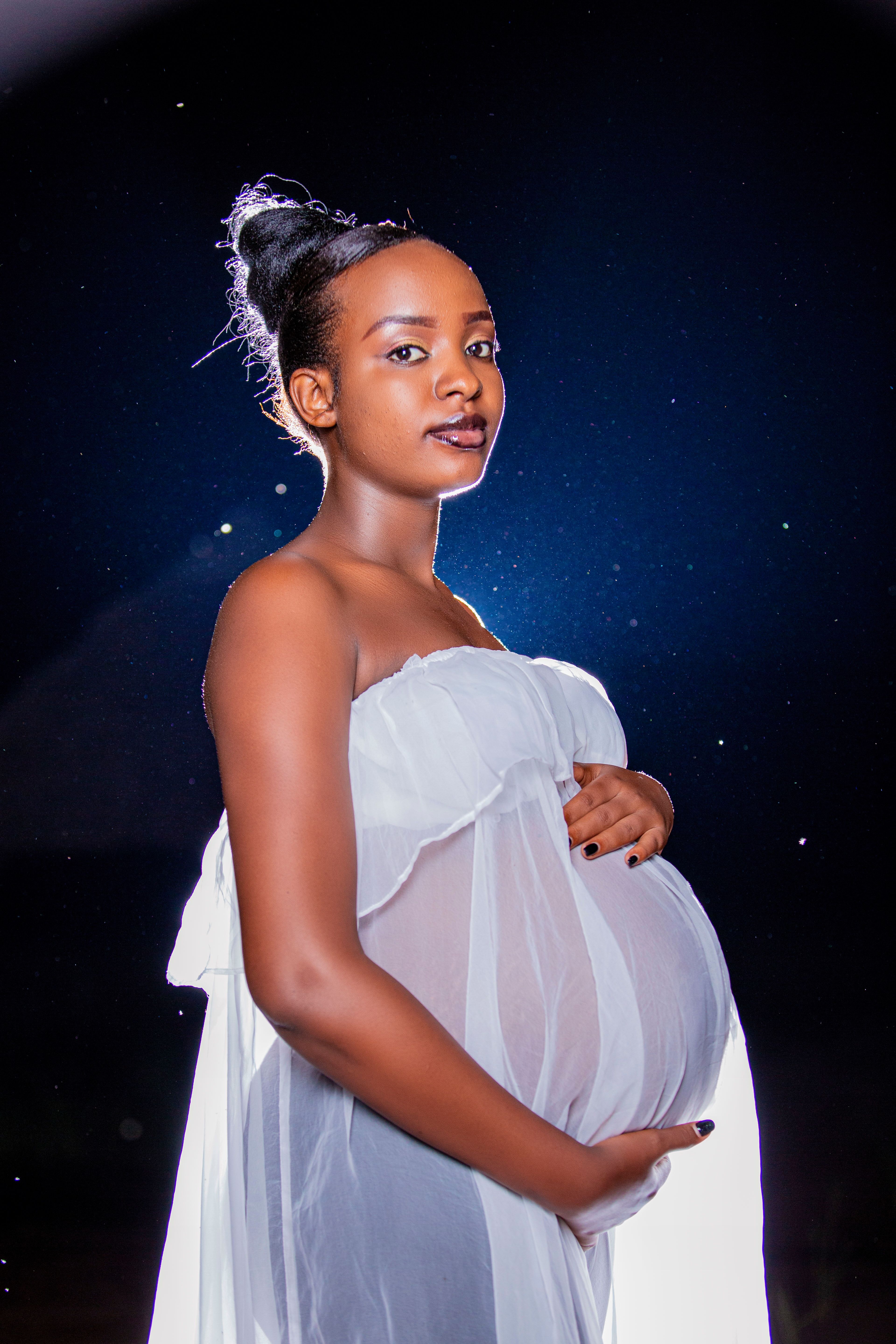 Pregnant woman poses gracefully in sheer white dress with starry backdrop.