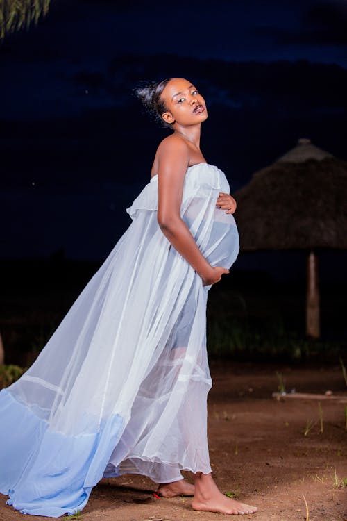 Free Barefooted Pregnant Woman in White Maxi Dress Holding Her Baby Bump while Posing at the Camera Stock Photo