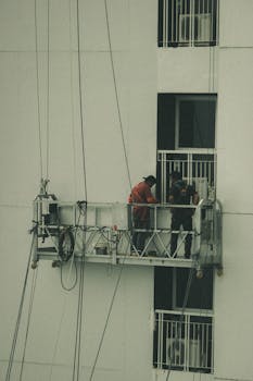 Two workers on a suspended platform engage in building maintenance work, providing an urban industrial scene.