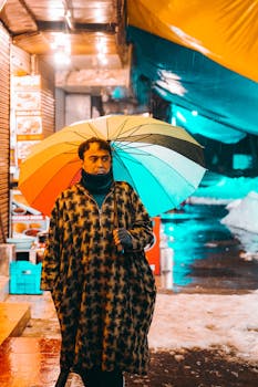 A man in a patterned coat walks under a colorful umbrella on a rainy urban street at night.