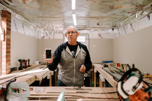 Senior man standing in a workshop, showcasing his smartphone with a focused expression.