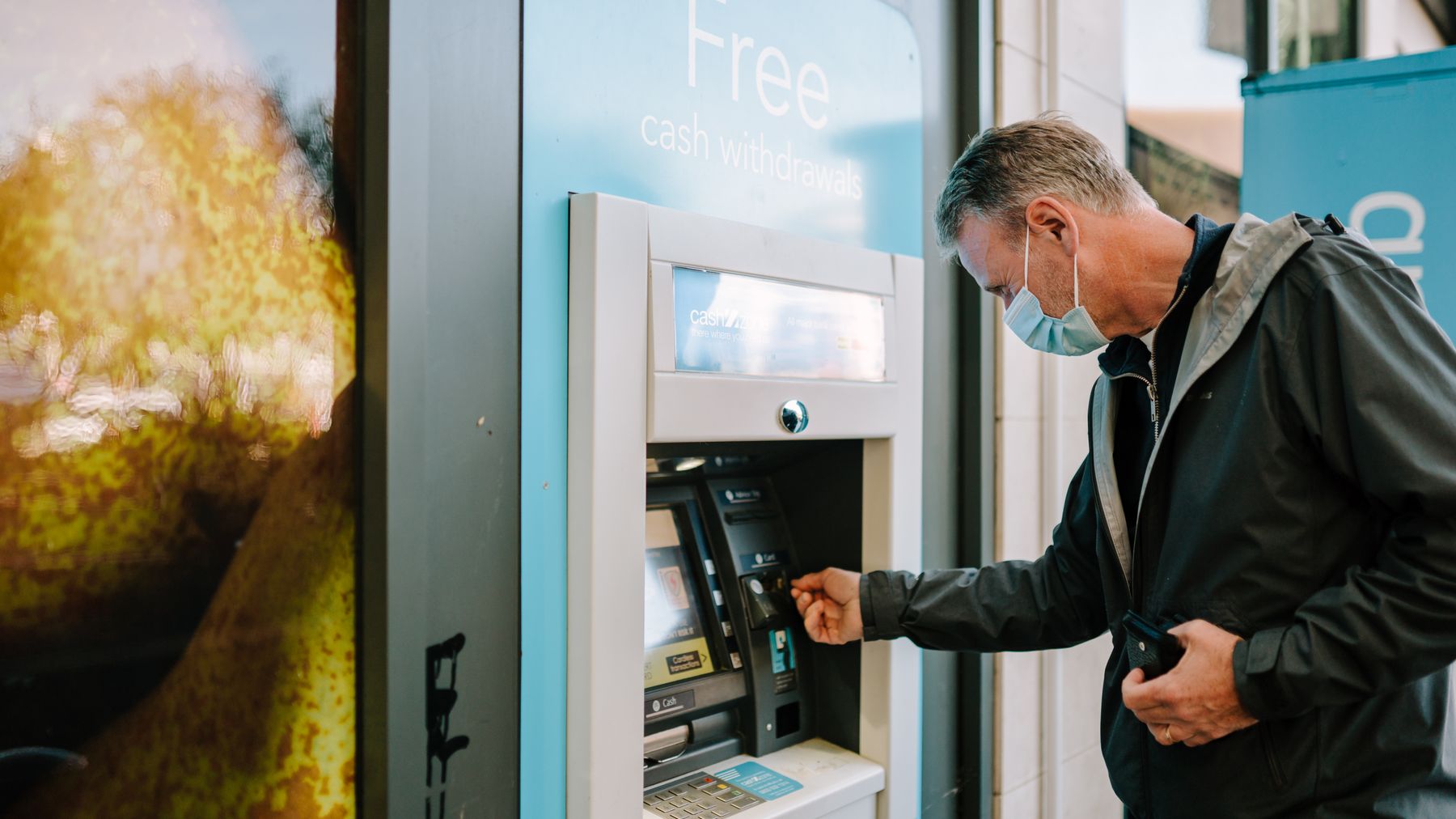 Một người đàn ông đeo khẩu trang đang sử dụng cây ATM ngoài trời để rút tiền mặt.