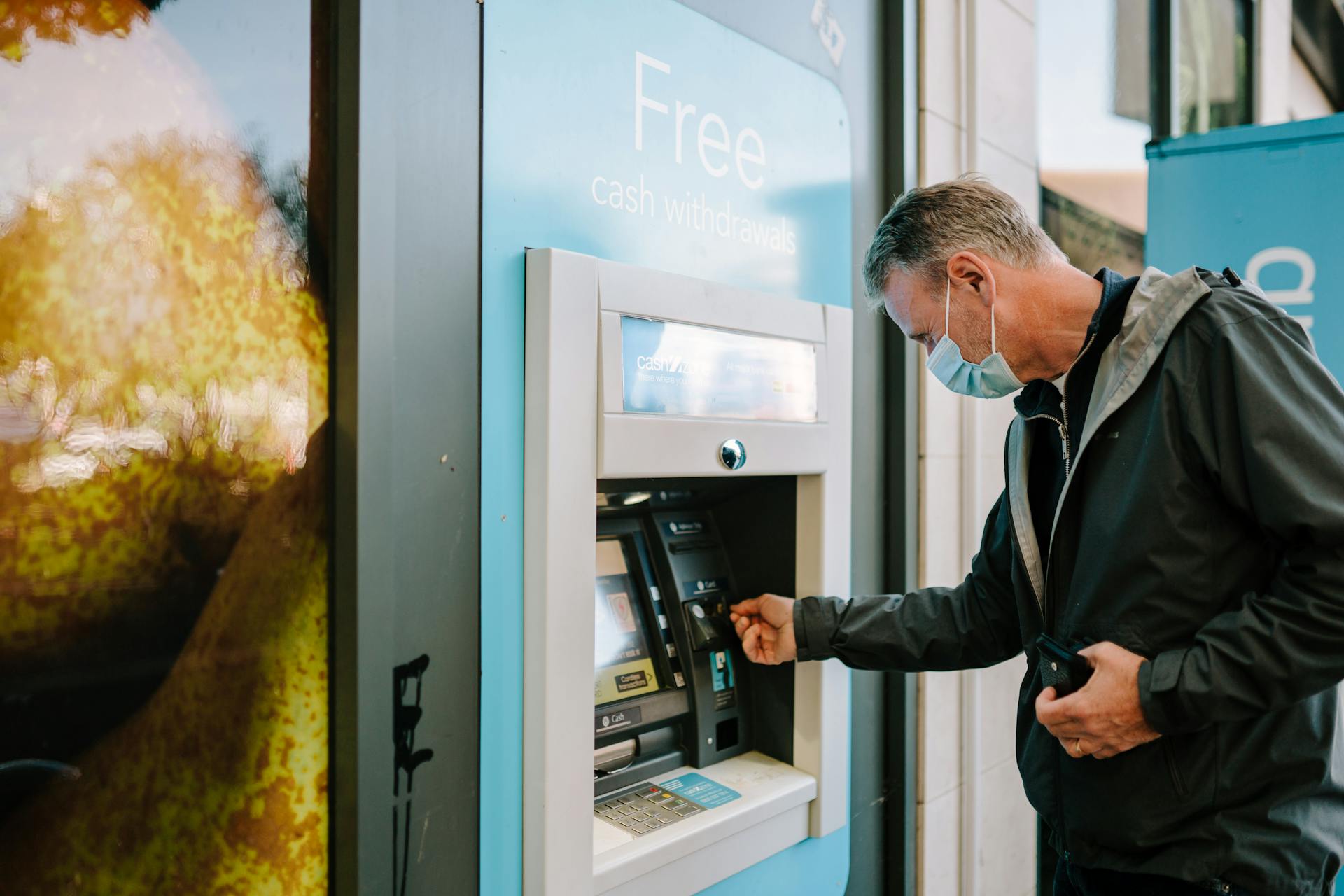 A man wearing a mask using an ATM machine outdoors for cash withdrawal.