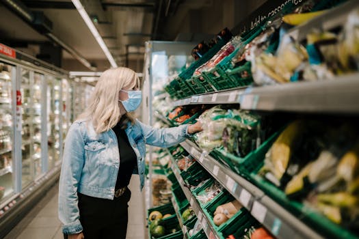 A woman in a mask shops for groceries in a supermarket aisle, selecting fresh produce.
