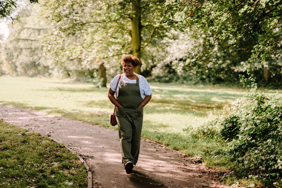 A joyful woman walks down a sunlit park path surrounded by lush greenery.