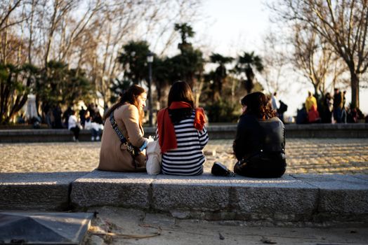 Three women sitting outdoors on a stone ledge, conversing in a sunny park.