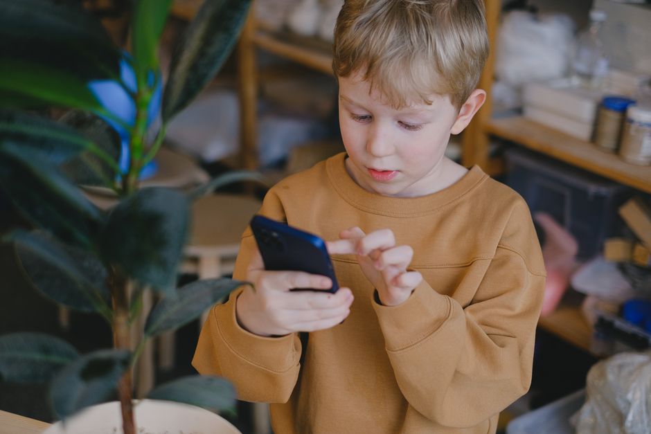 Ein kleiner Junge in einem braunen Pullover benutzt ein Smartphone in der Nähe einer Topfpflanze im Haus.