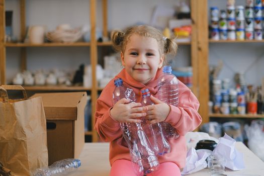 Young girl in pink sweater holding plastic bottles, advocating recycling indoors.